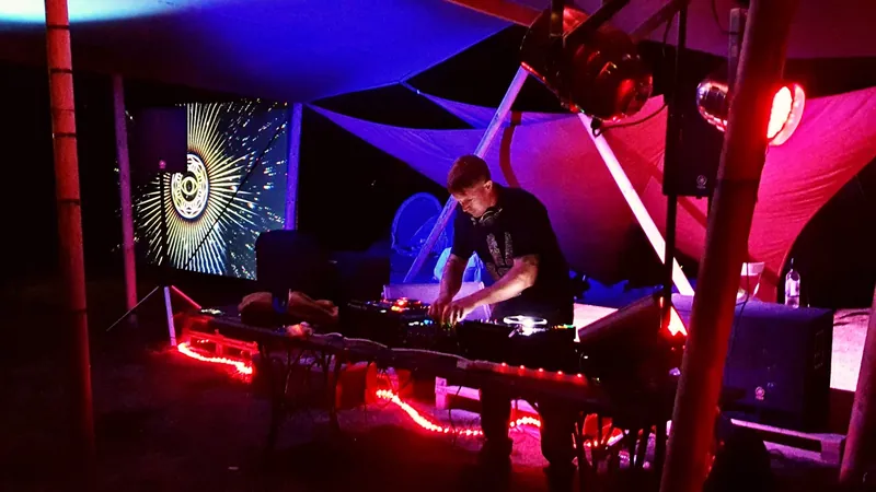Scruff Baggins DJing under a canopy at night, lit red and blue, with visuals on a screen behind