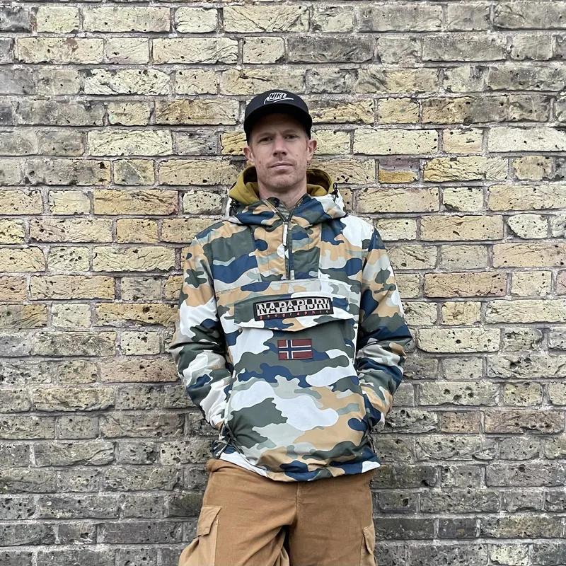 Portrait of Scruff Baggins in a camo jacket and cap against a London brick wall