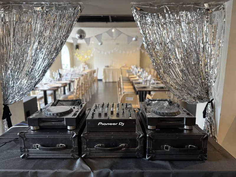 Pioneer decks and mixer set up for a wedding, framed by silver tinsel curtains, with the dining room laid out behind