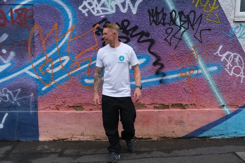 Scruff Baggins in a white T-shirt in front of a colourful graffiti wall